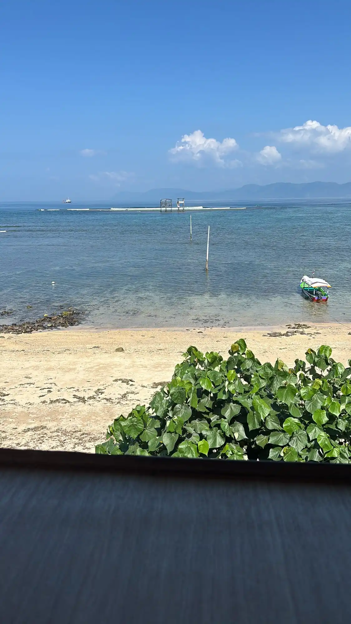 View from a beachfront restaurant looking out over turquoise water with local boats anchored near the reef, mountains visible in the background