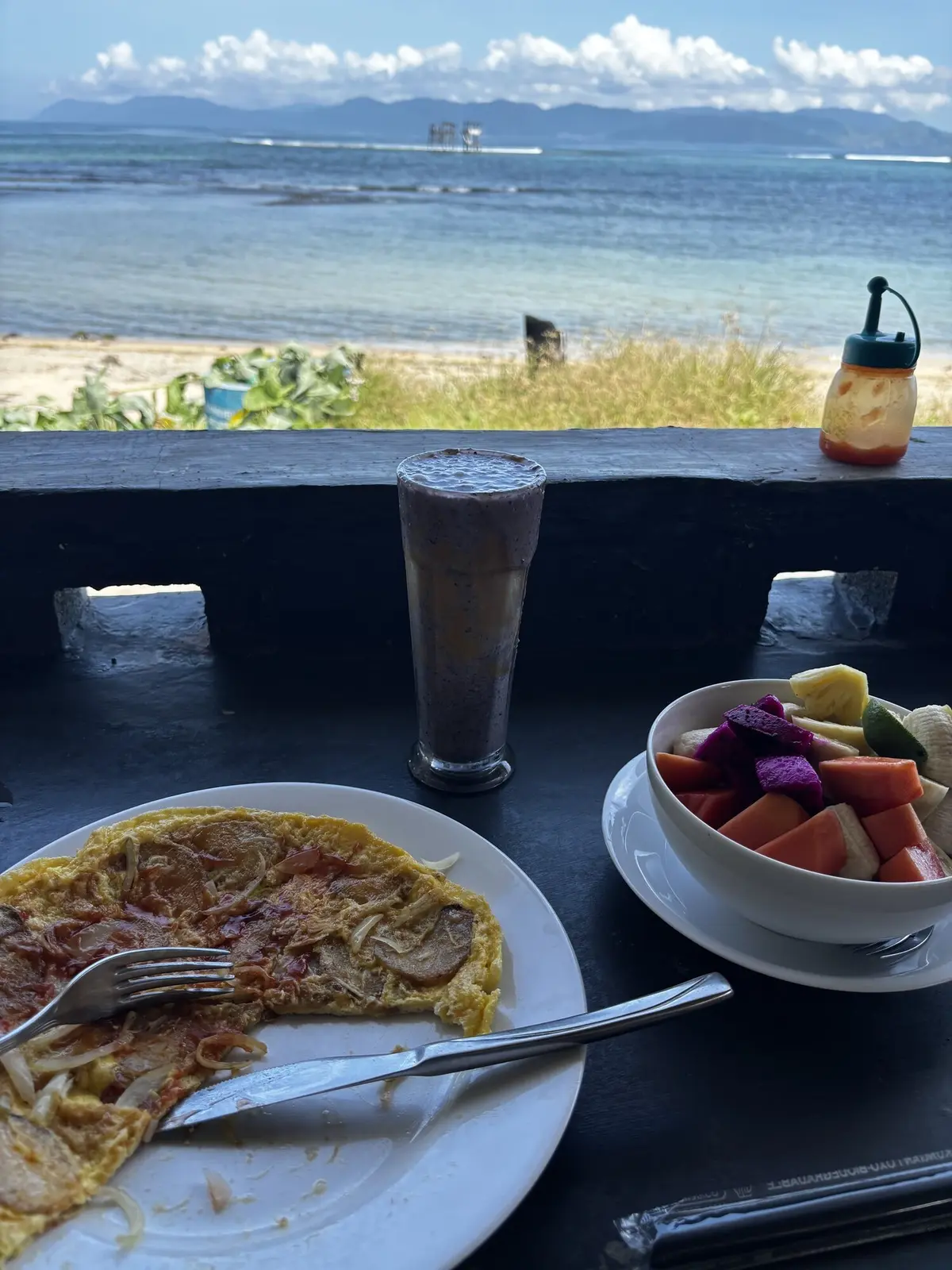Breakfast spread of omelette, tropical fruit bowl and smoothie at the beachfront cafe, ocean and boats visible through the railing