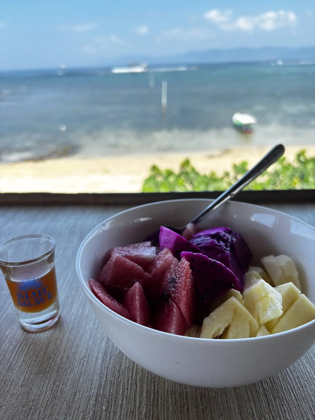Fresh fruit bowl loaded with dragon fruit, watermelon, pineapple and banana at a beachfront cafe, turquoise ocean in the background