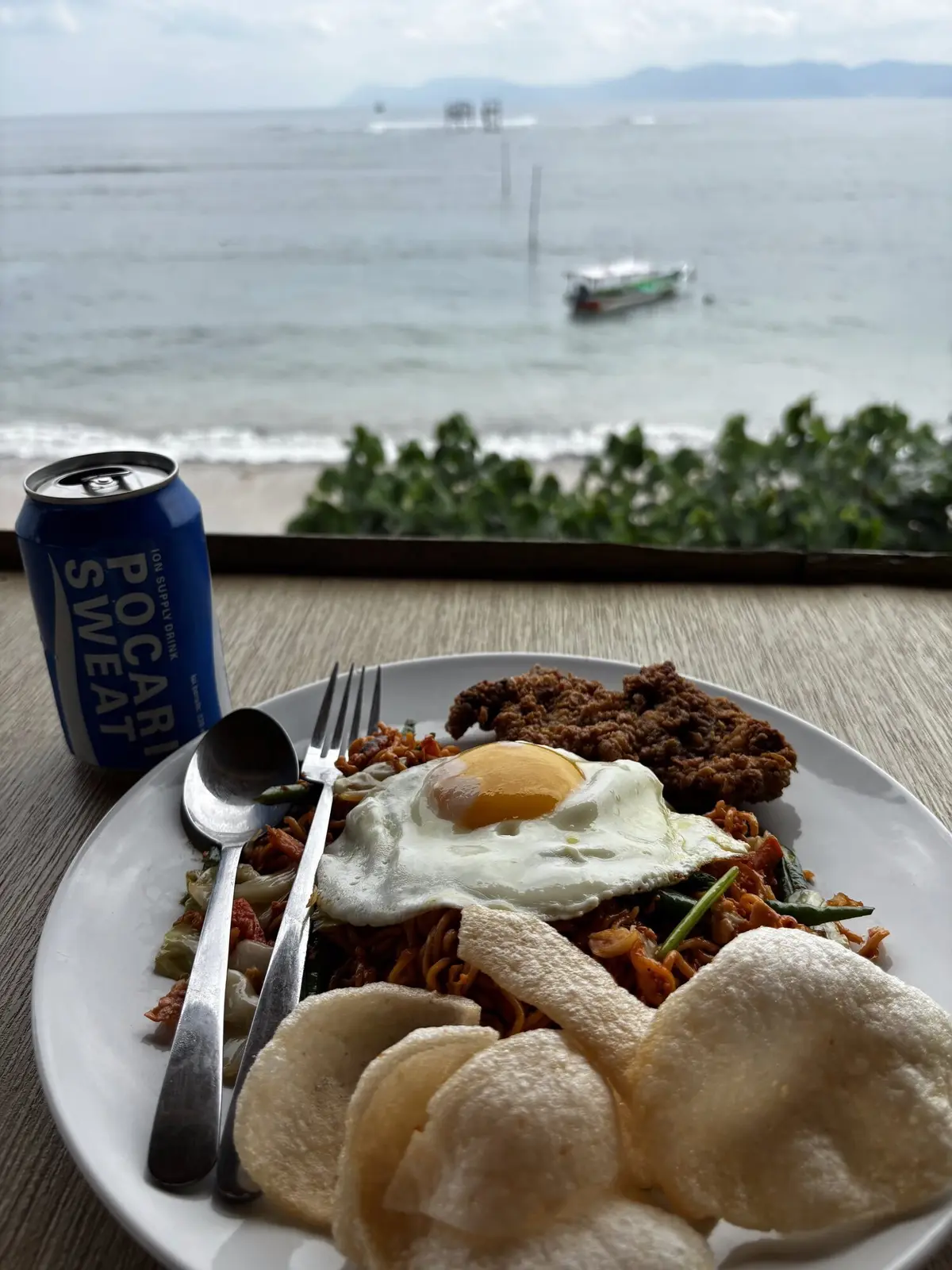 Plate of nasi goreng with fried egg and prawn crackers alongside a Pocari Sweat can, ocean and boats visible in the background from the beachfront cafe