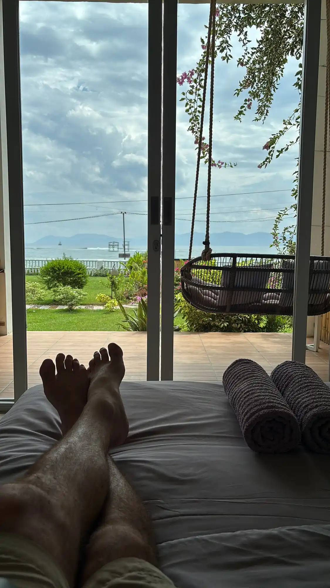 View from the bed at Peak Surf House looking out through glass doors to the ocean, with a patio swing chair and garden in between