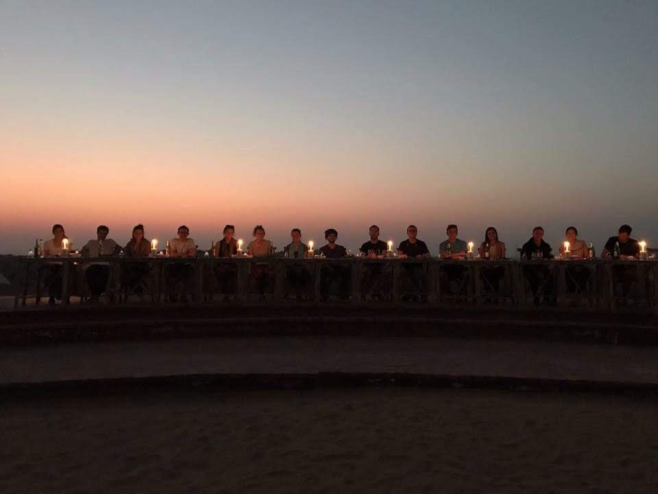 Long candlelit dinner table in the Thar Desert at sunset with fellow travellers silhouetted against the sky