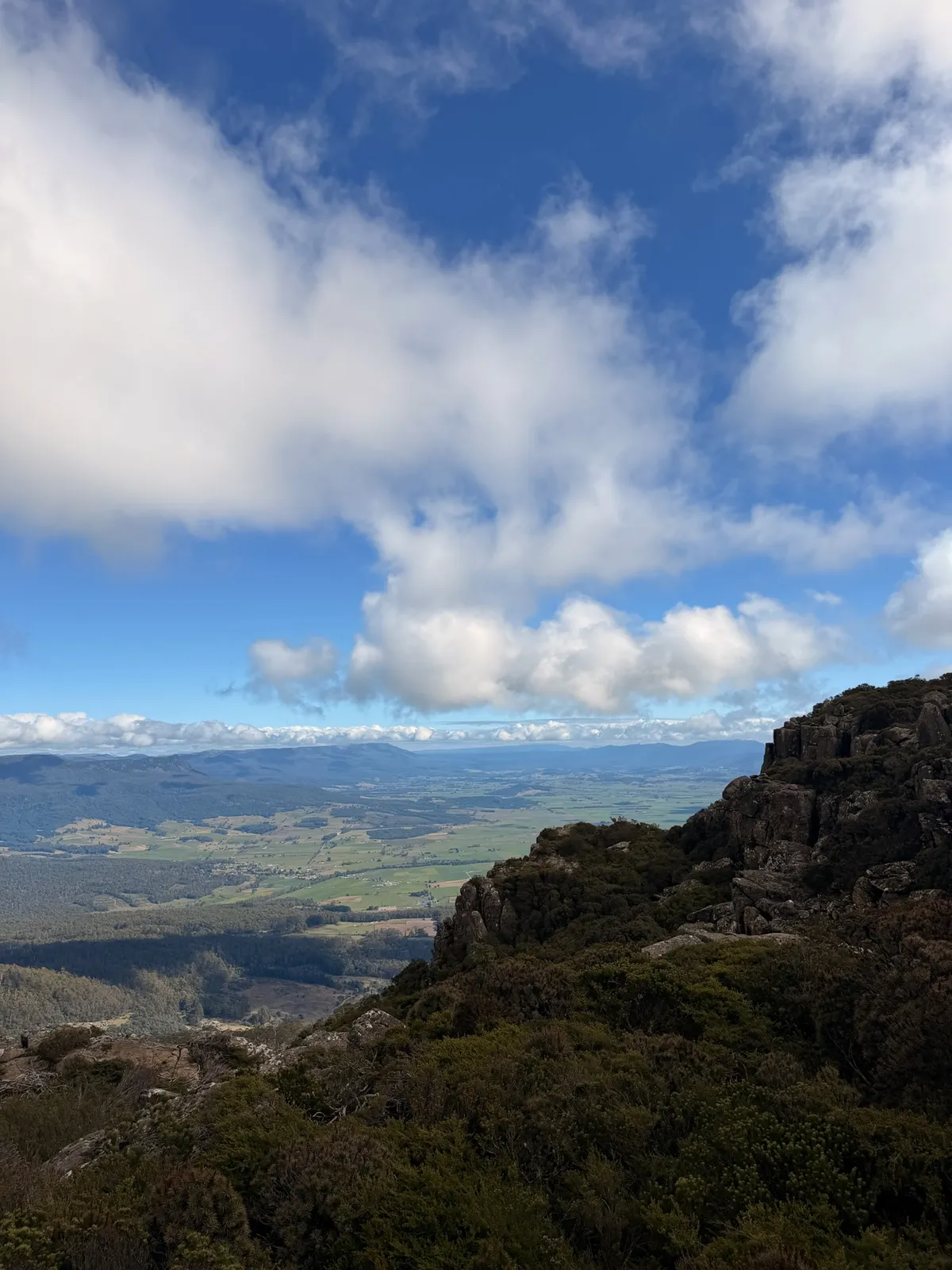 Summit view from Quamby Bluff showing dolerite rock outcrops, alpine scrub, and green farmland stretching to distant ranges under broken cloud